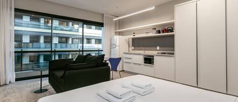 A modern hotel room with a large bed, a sofa, a small table, and a kitchen area.