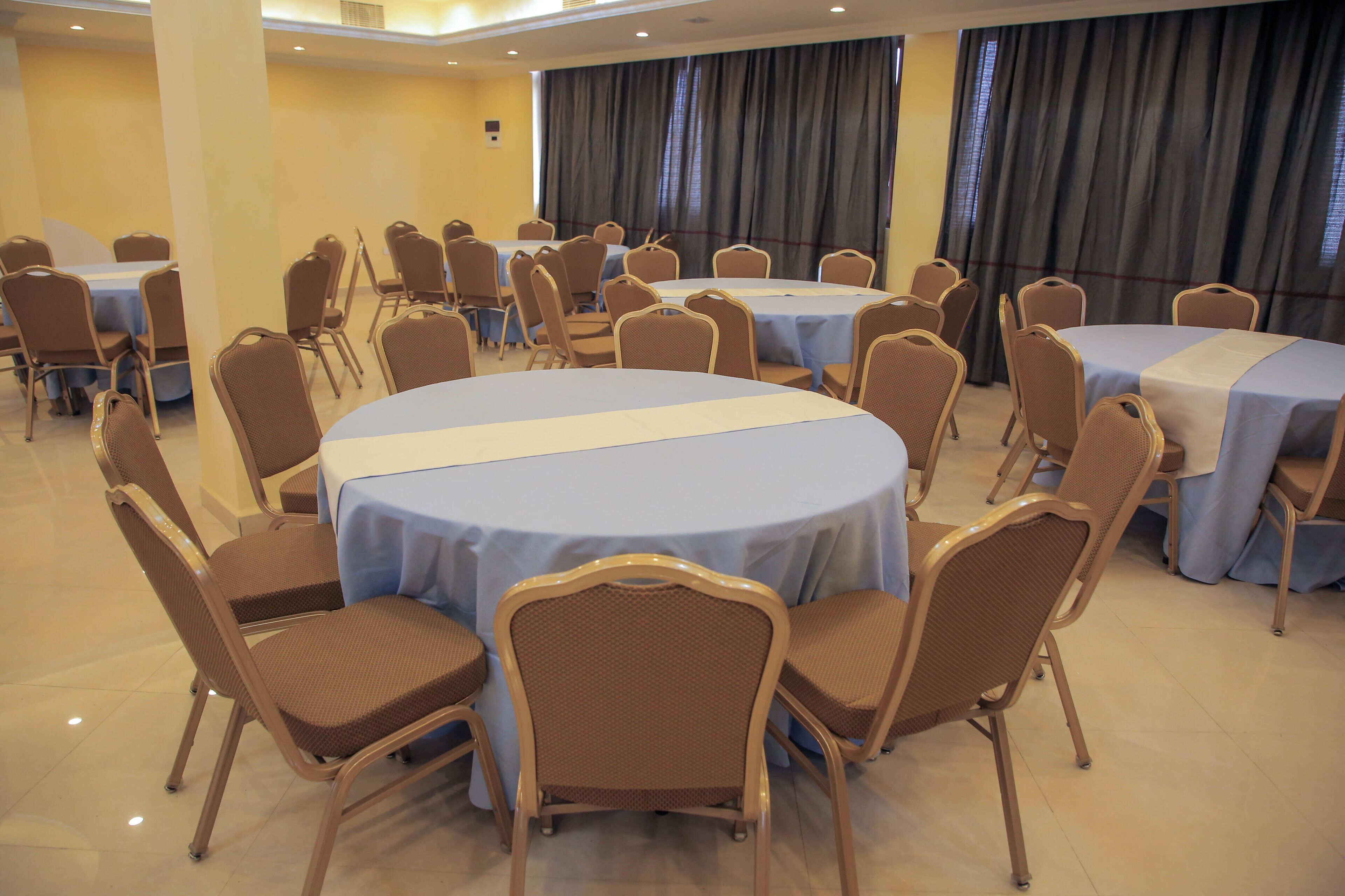 Un salón de conferencias con mesas y sillas redondas, amplias ventanas con cortinas y una pared amarilla.