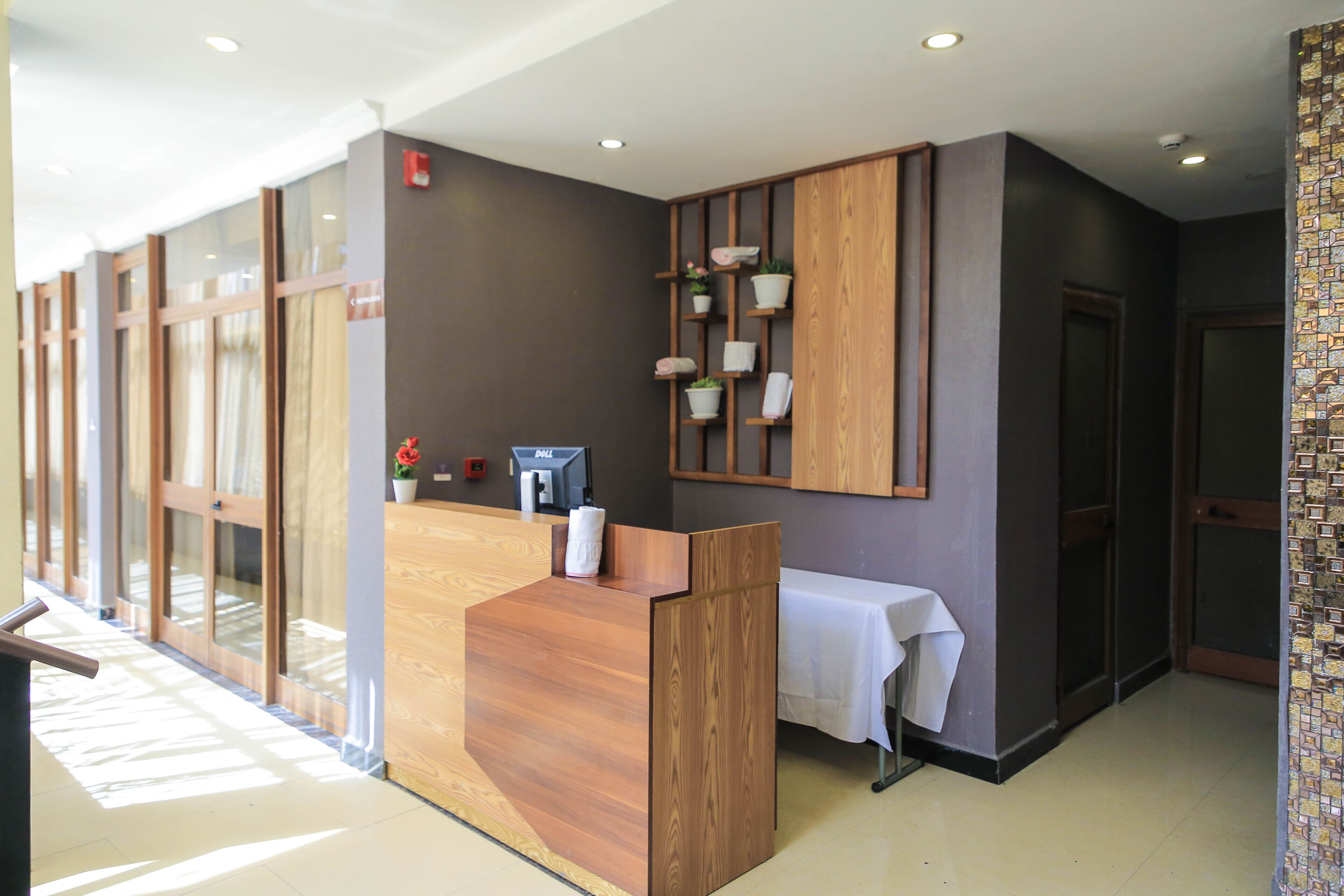A reception area with a wooden reception desk, a small table with a white tablecloth, and a wall-mounted shelf with potted plants and towels.
