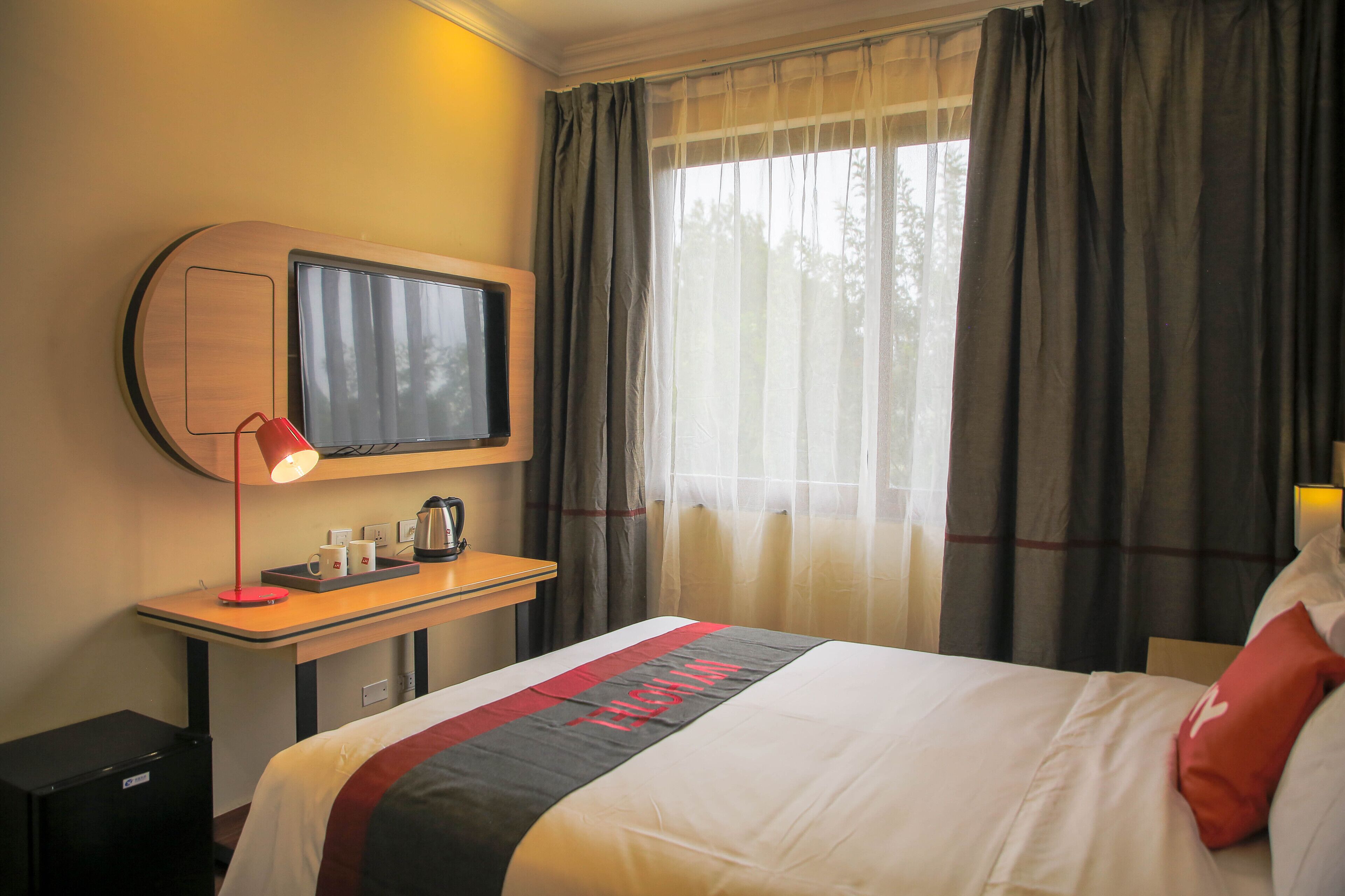 Een hotelkamer met een bed, een bureau met lamp, een televisie en een raam met gordijnen.