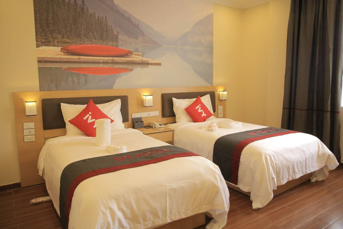 A hotel room with two beds, each with a red pillow and a white blanket. There is a painting of a lake and mountains on the wall above the beds.