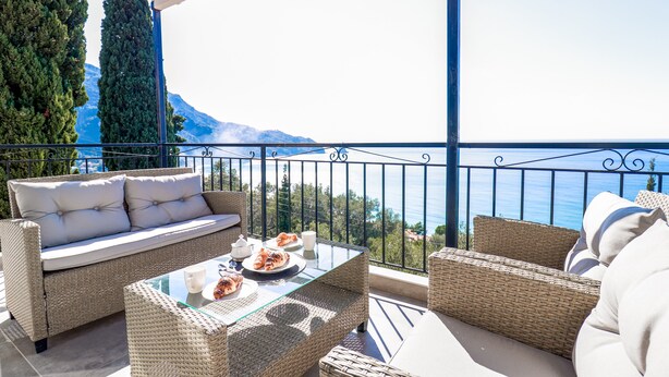 A balcony with wicker furniture, a glass table, and a view of the sea and mountains. - Maison du Soleil by CorfuEscapes (Corfu)