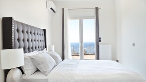 A modern bedroom with a large bed, a nightstand, a lamp, and a view of the sea through a sliding glass door. - Maison du Soleil by CorfuEscapes (Corfu)