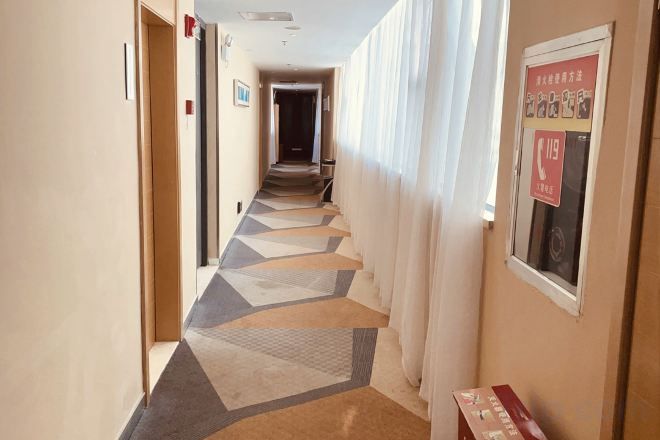 En hotellkorridor med ett mönstrat golv, en dörr till vänster, en affisch till höger och ett fönster med gardiner.