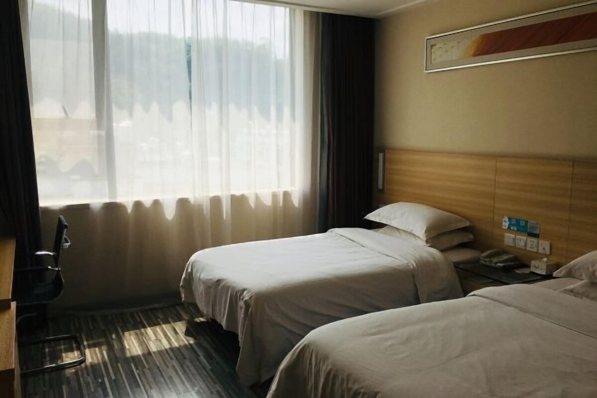 Ein Hotelzimmer mit zwei Betten, einem Holz-Kopfteil, einem Fenster mit durchscheinenden Vorhängen, einem Schreibtisch mit Stuhl und einem gerahmten Bild an der Wand.
