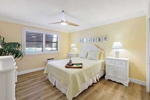 A bedroom with a bed, bedside tables, a ceiling fan, a window with blinds, and wall-mounted artwork. - Rare Garden Unit | Pool, Grills & Beach Access (Marco Island)