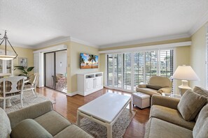 A living room with a white coffee table, beige sofas, a white cabinet, and a large window with blinds. - Rare Garden Unit | Pool, Grills & Beach Access (Marco Island)