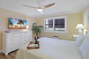 A bedroom with a bed, a ceiling fan, a TV, a window with blinds, a bedside lamp, and a plant. - Rare Garden Unit | Pool, Grills & Beach Access (Marco Island)