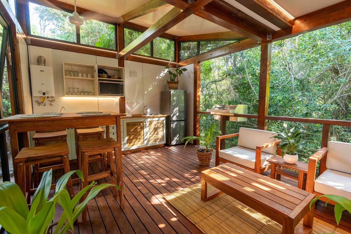 Eine Holzterrasse mit Küchenbereich, inklusive Kühlschrank, Spüle und Regalen mit Gläsern. Holzstühle und ein Tisch mit Kissen sind ebenfalls vorhanden.