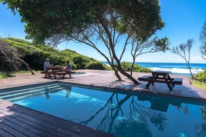 Área de piscina com deck de madeira, mesas para piquenique e vista para o oceano.