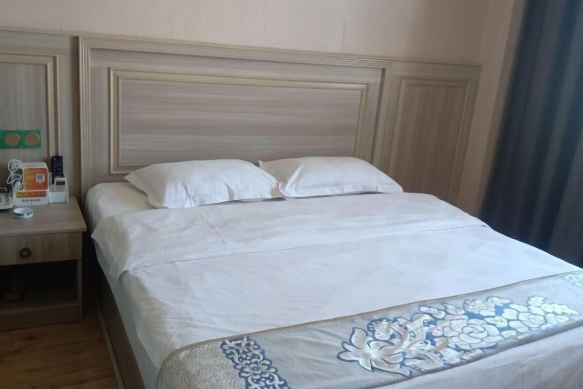 A neatly made bed with a floral-patterned bedspread, a wooden headboard, and a nightstand with a remote control and a box.