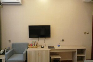 A room with a flat-screen TV mounted on the wall, a light blue armchair, a wooden desk with a laptop, and a small shelf. - Chuxiong Haiheng Hotel (Chuxiong City)