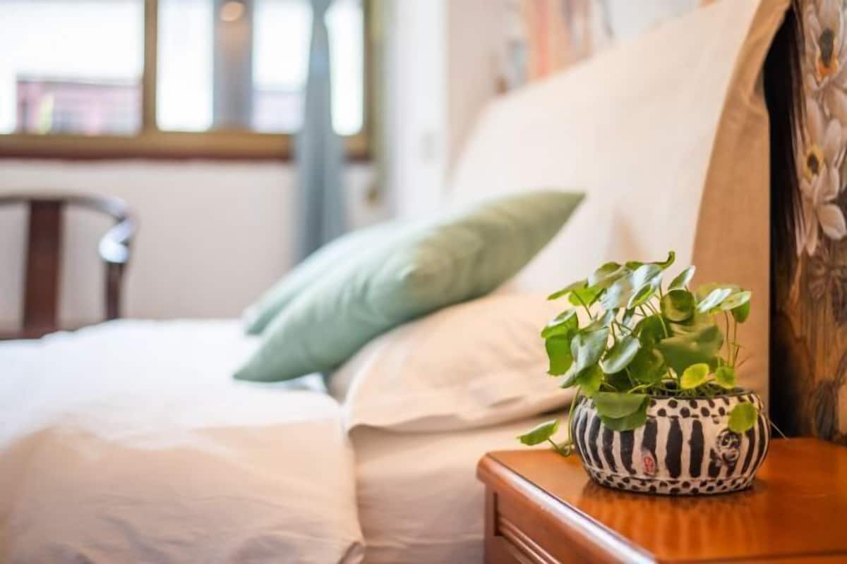 Una cama con un almohadón verde, una mesita de noche de madera con una maceta a rayas y una ventana al fondo.