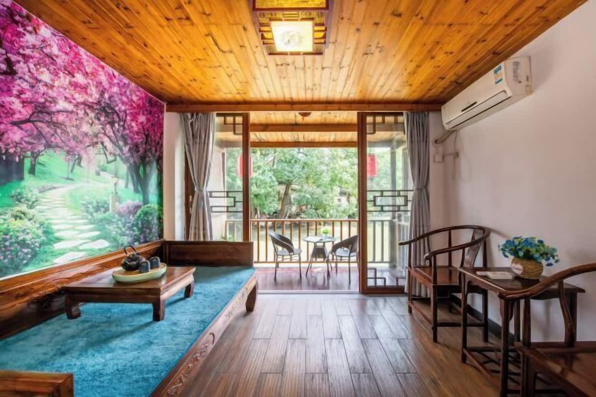 Una habitación con techo de madera, un cuadro grande de un jardín, un banco de madera, una mesa con una bandeja y vistas a un balcón exterior a través de una puerta de cristal.