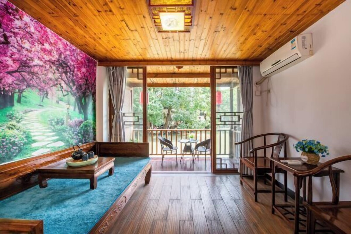 A wooden-floored room with a large window, a wooden bench, a table with a teapot, and a view of a garden.
