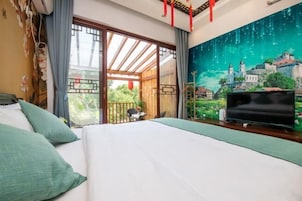 Una habitación de hotel con una cama grande, televisión, balcón con vistas a zonas verdes y un mural de un pueblo en la pared.