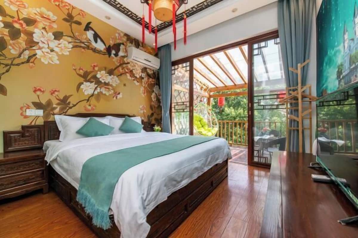 Un dormitorio con cama de madera, almohadas verdes, un mural floral y un ventanal con vistas a la vegetación.