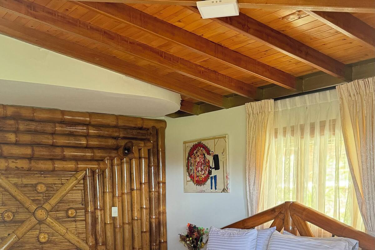 A wooden-framed bedroom with a bed, a wooden ceiling, a wall-mounted light fixture, and a window with curtains.