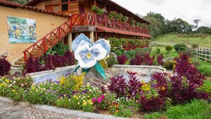 A wooden building with a red staircase and balcony, surrounded by colorful flowers and a large flower sculpture. - Jardín de Silleteros Agro Parque Hotel (Envigado)