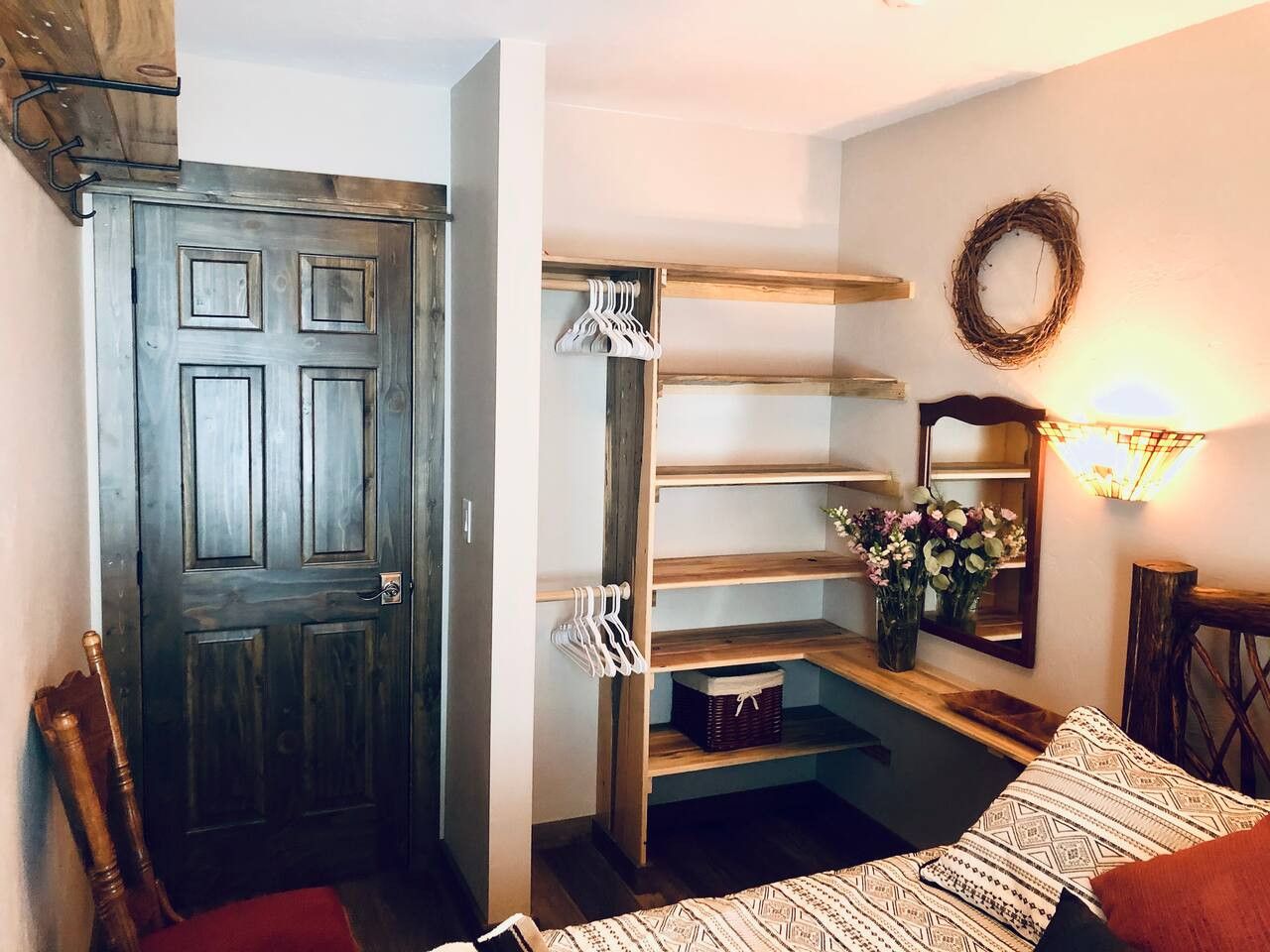 A room with a wooden door, a built-in shelving unit with hanging clothes, a mirror, and a wall-mounted light fixture.