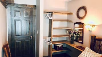 A room with a wooden door, a built-in shelving unit with hanging clothes, a mirror, and a wall-mounted light fixture.
