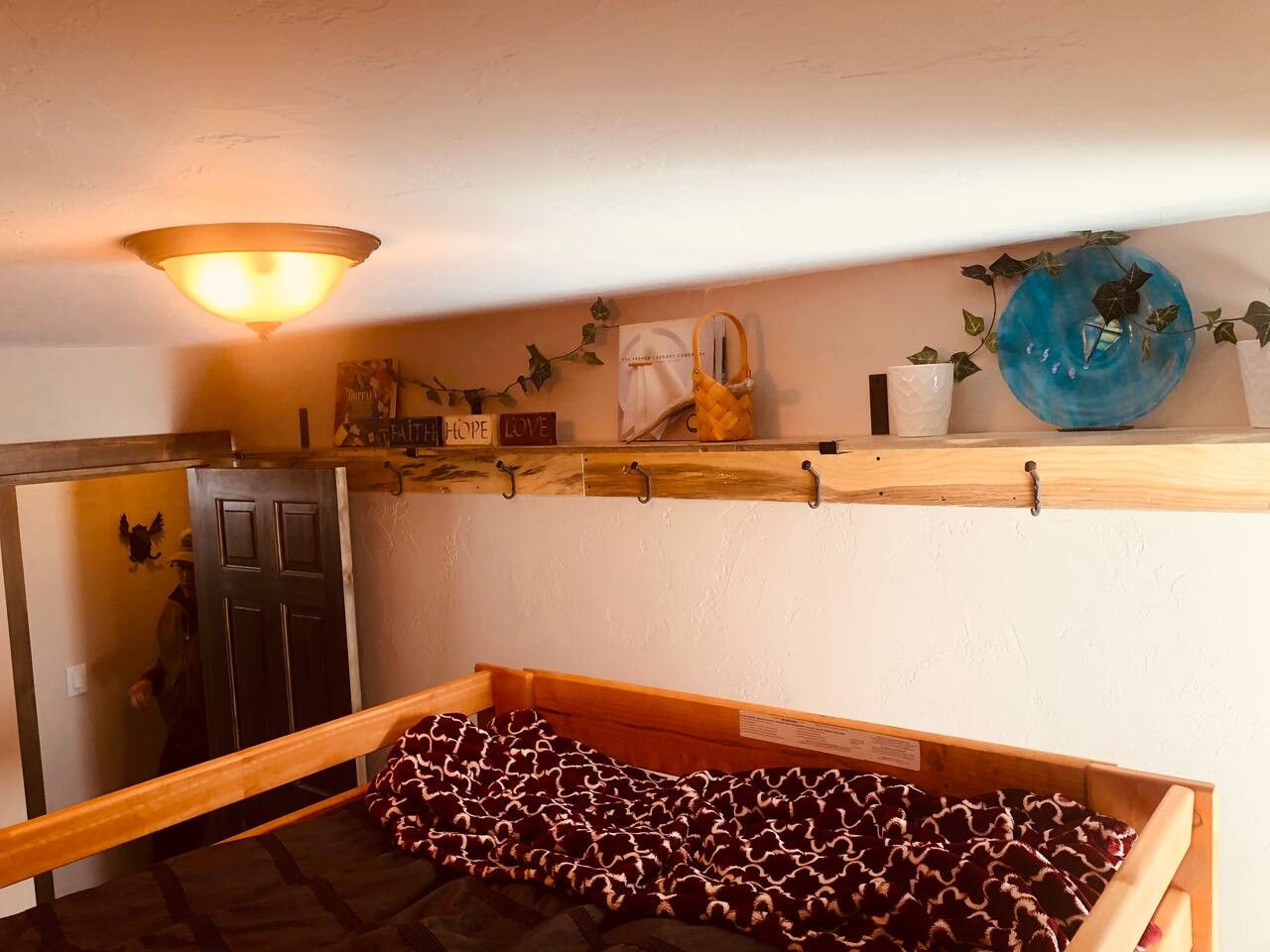 Una habitación con una cama, un estante de madera con diversos objetos, una lámpara de pared y una puerta.