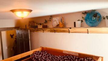 Una habitación con una cama, un estante de madera con diversos objetos, una lámpara de pared y una puerta.
