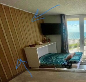 Una habitación con cama, televisor montado en la pared, una mesita con adornos y vista al mar desde un balcón.