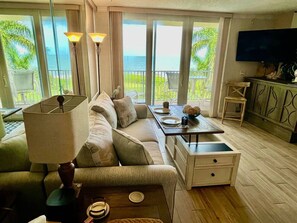 A living room with a sofa, a coffee table, a TV, and a balcony with a view of the ocean.