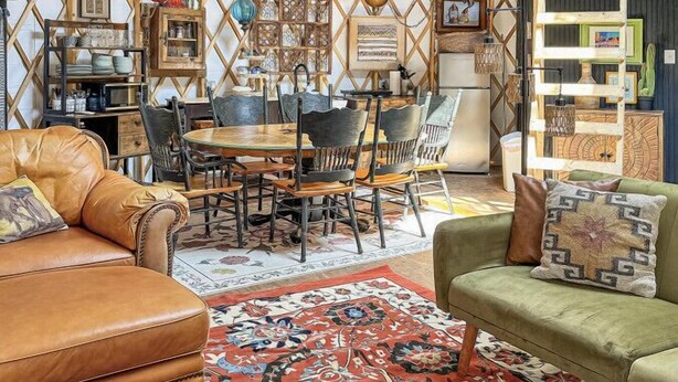Un interior acogedor y bien iluminado, con techo alto, un comedor con mesa y sillas de madera, y una zona de estar con sofá y sillones.