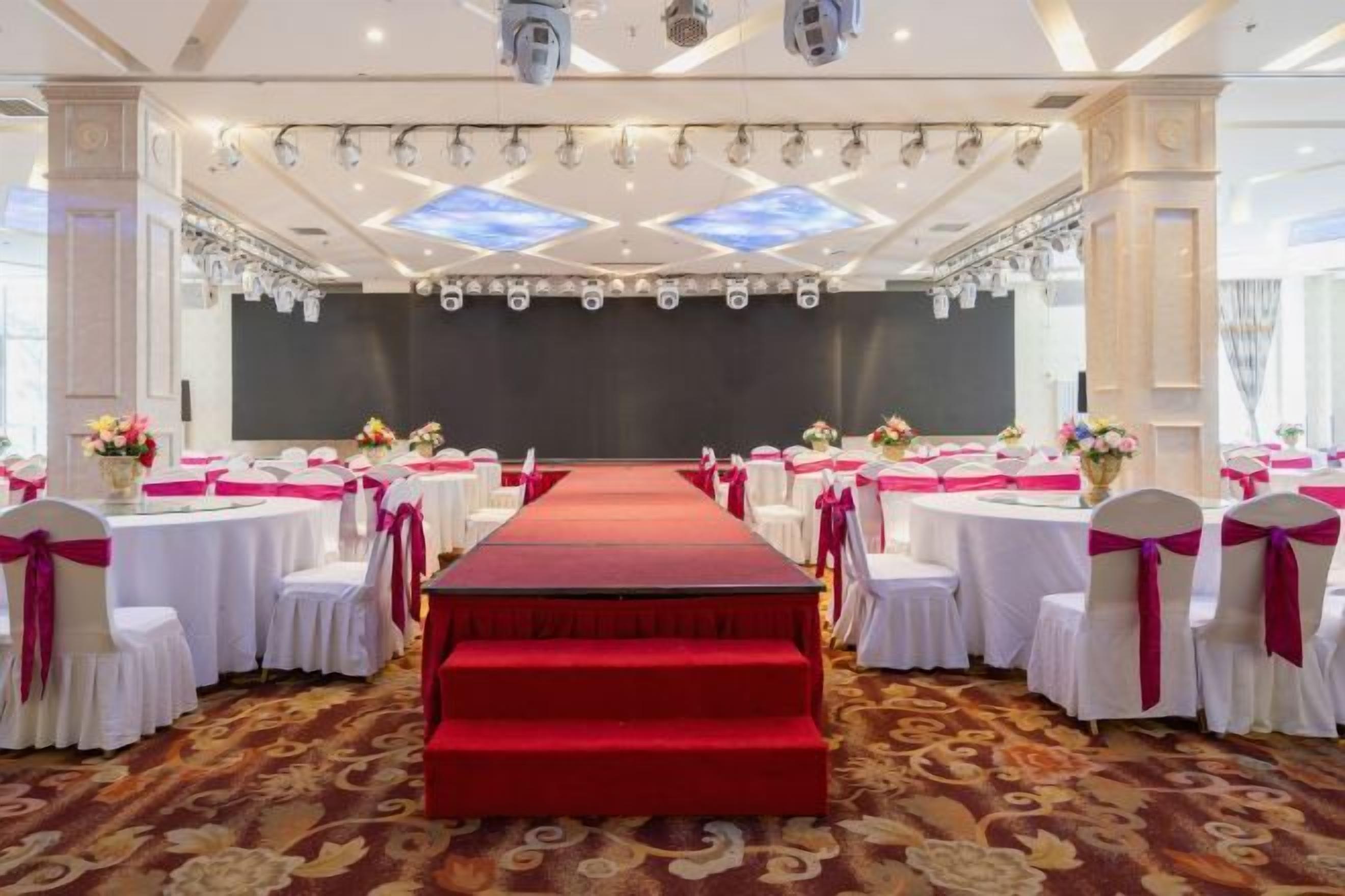 A banquet hall with a central stage, rows of round tables with white tablecloths and pink sashes, and a red carpet leading to the stage.