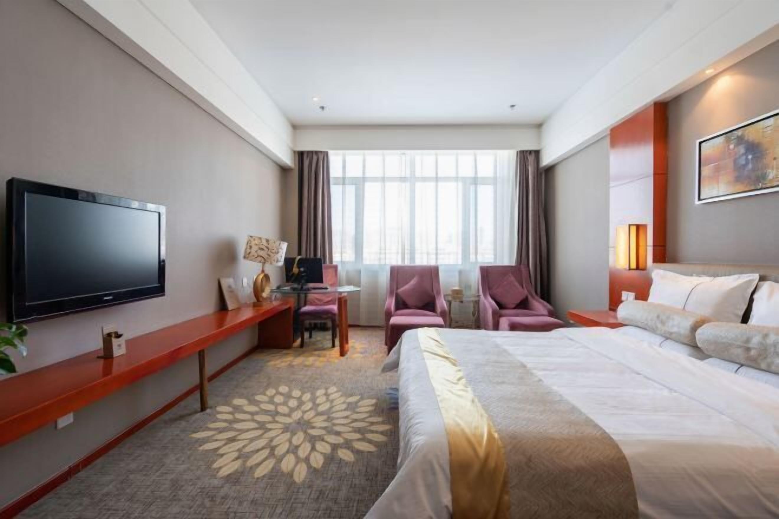 Quarto de hotel com cama, escrivaninha com televisão de tela plana, cadeira, luminária de mesa e janela com cortinas.