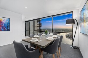Un espace de salle à manger moderne, avec une grande table dressée pour six personnes, de grandes fenêtres offrant une vue sur la ville et un lampadaire.