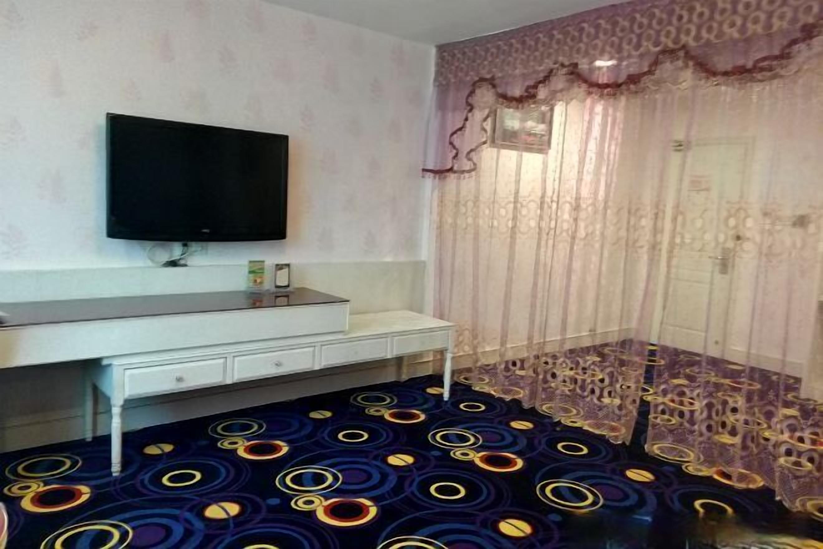 A room with a flat-screen TV mounted on the wall, a white desk with drawers, a patterned carpet, and a window with sheer curtains.