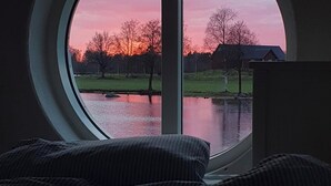 A round window with a view of a sunset over a body of water and trees.