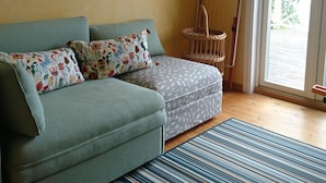 Un salon comprenant un canapé, des coussins à motifs, un tapis à rayures, un fauteuil en osier et une porte-fenêtre coulissante donnant sur un espace extérieur.