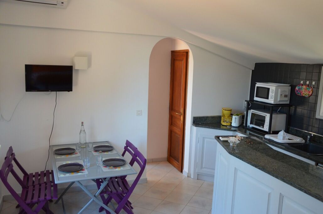 A compact kitchen with white cabinets, a microwave, and a toaster. There is a dining table with purple chairs and a flat-screen TV mounted on the wall.