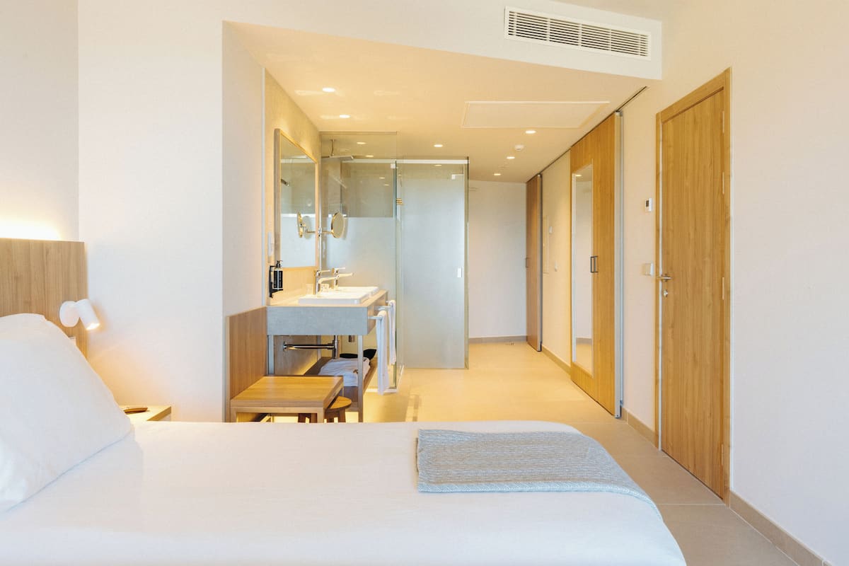 A hotel room with a bed, a bedside table, a nightstand, a bathroom with a sink and mirror, and a wooden wardrobe.