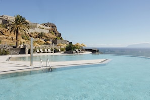 A clear swimming pool with lounge chairs and umbrellas, a stone wall, and a view of the ocean.