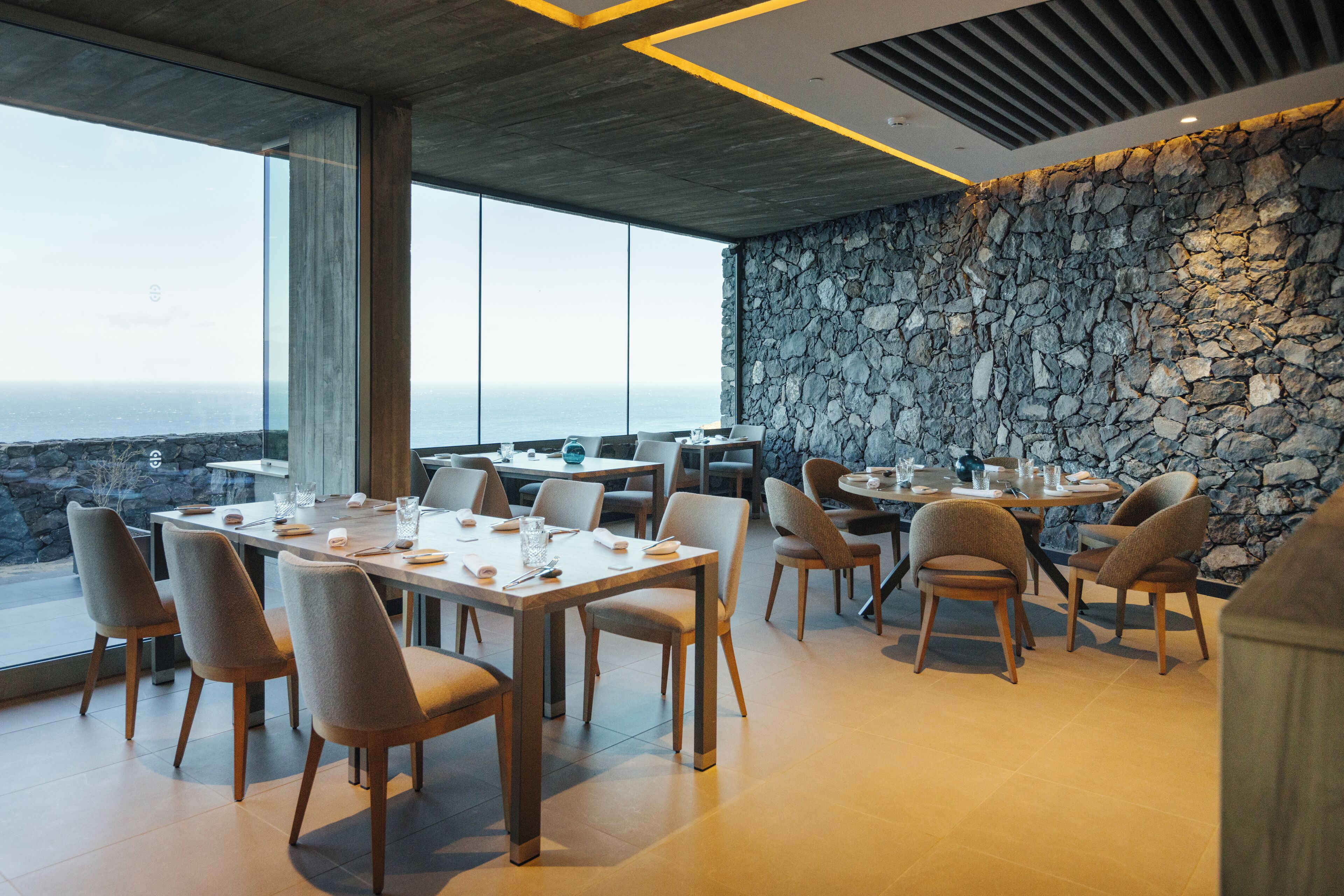 A modern dining area with a stone wall and large windows overlooking the ocean.