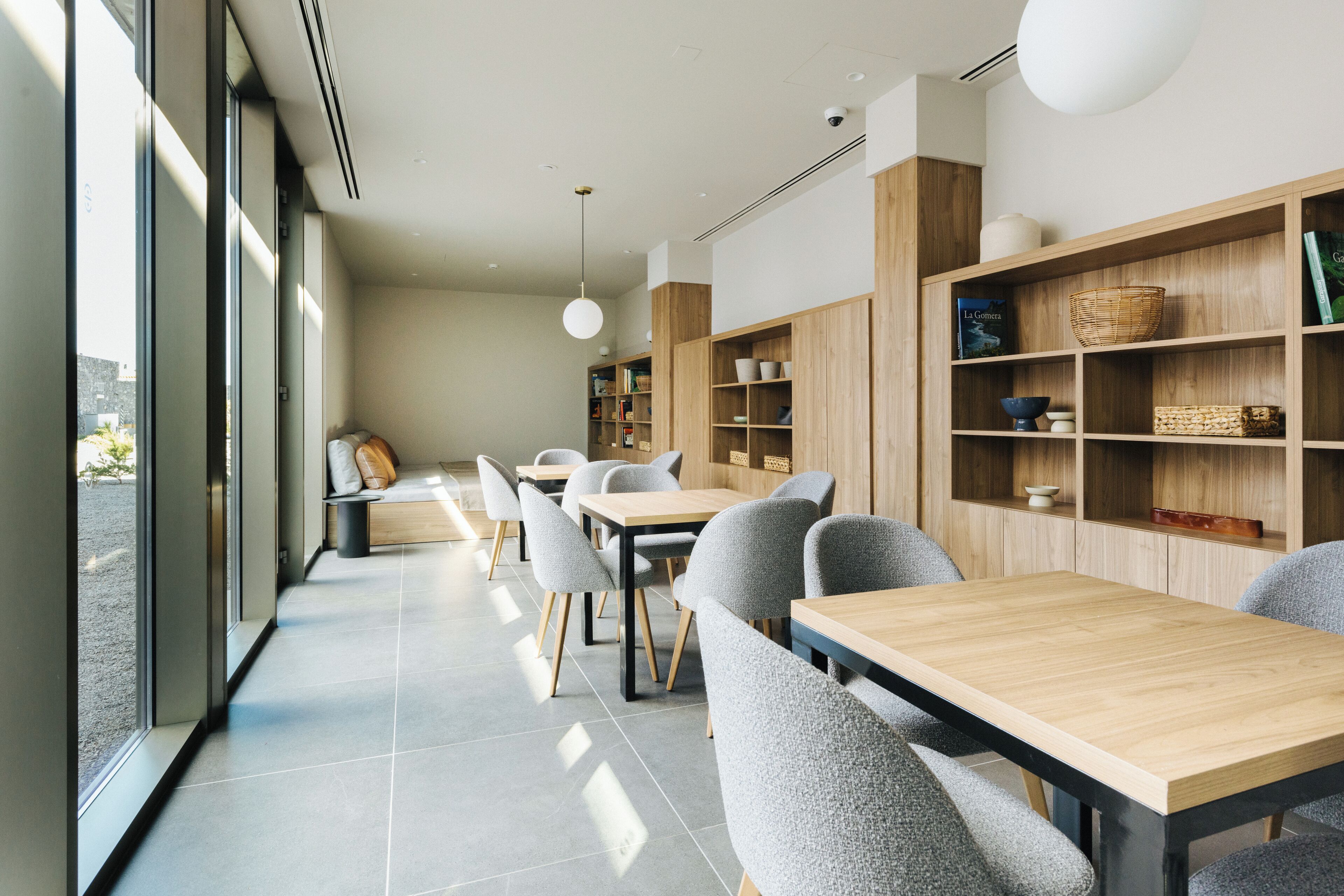 A modern dining area with wooden tables and grey upholstered chairs, large windows, and built-in wooden shelves with various items.
