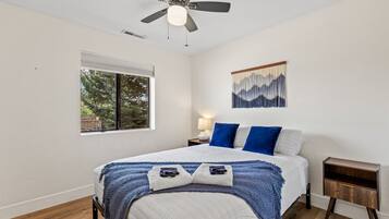 Une chambre à coucher comprenant un lit, un ventilateur de plafond, une fenêtre, une table de chevet et une œuvre d’art accrochée au mur.