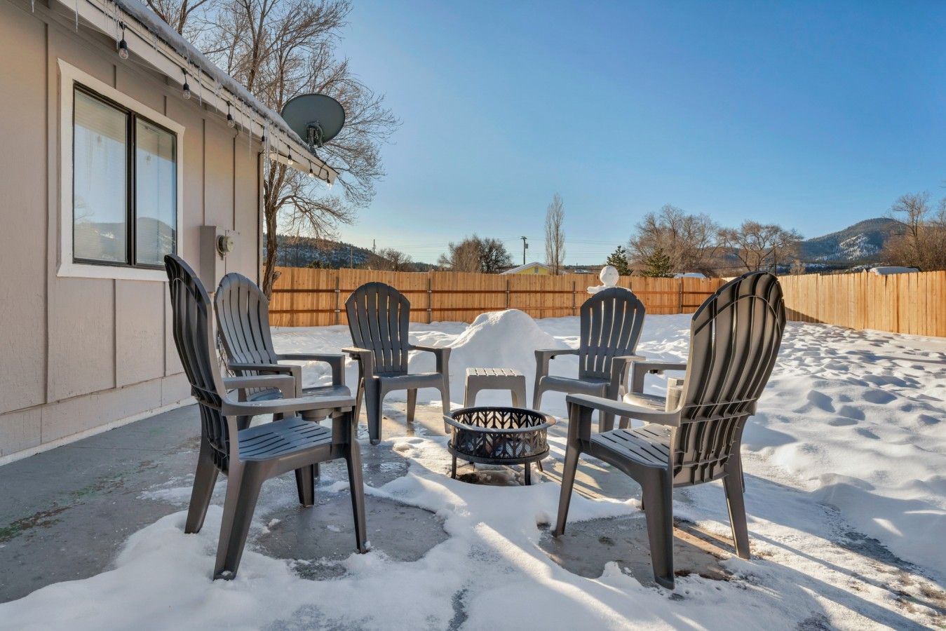 A patio with four chairs and a table, surrounded by snow, with a wooden fence and a satellite dish in the background.