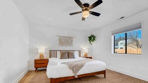 Un dormitorio con una cama grande, cabecera de madera, dos mesitas de noche con lámparas, un ventilador de techo, una ventana con vistas y una planta colgante.