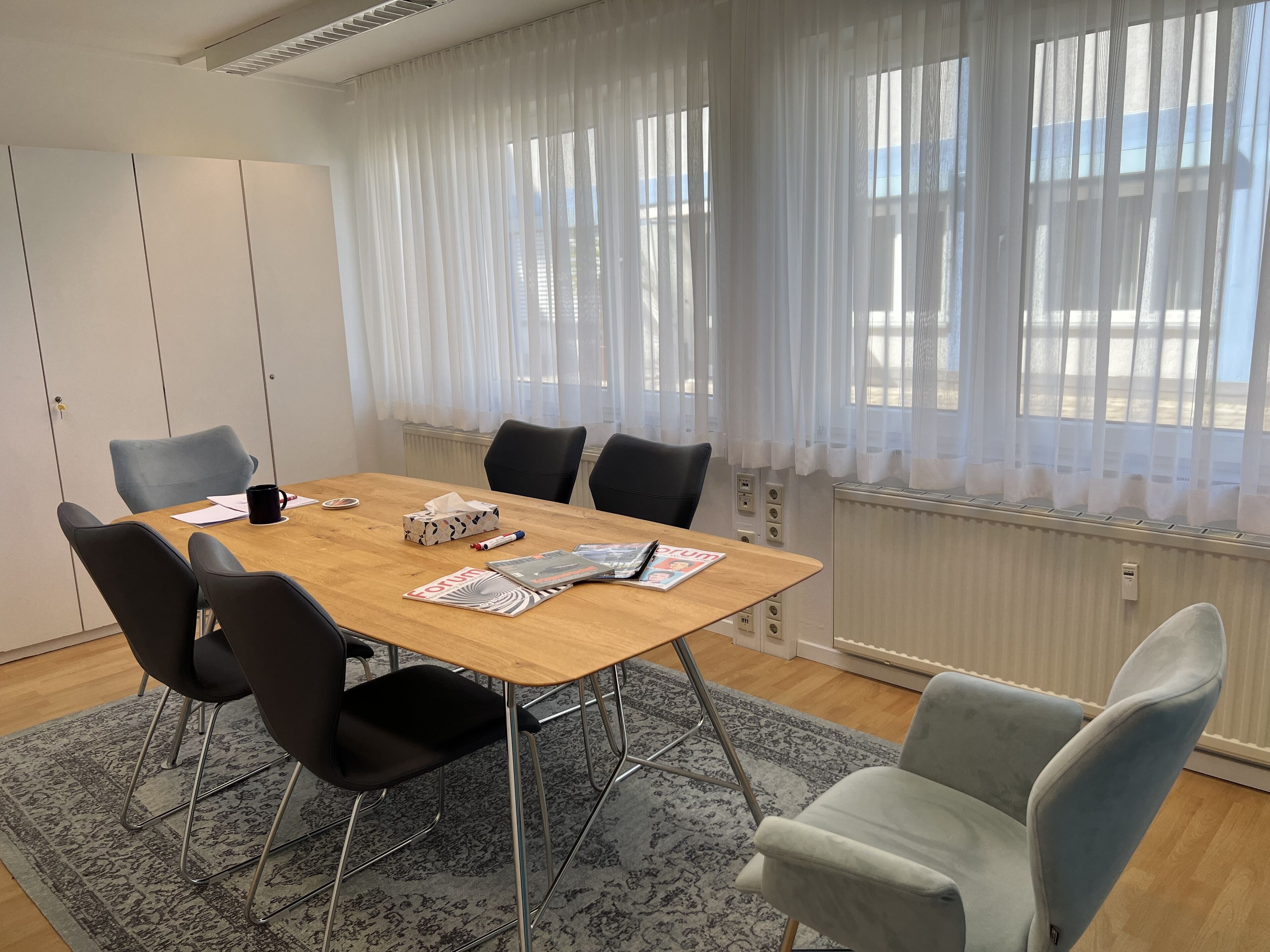 Uma sala de conferências com mesa de madeira, cadeiras pretas e cinzentas, tapete estampado e janelas amplas com cortinas translúcidas.