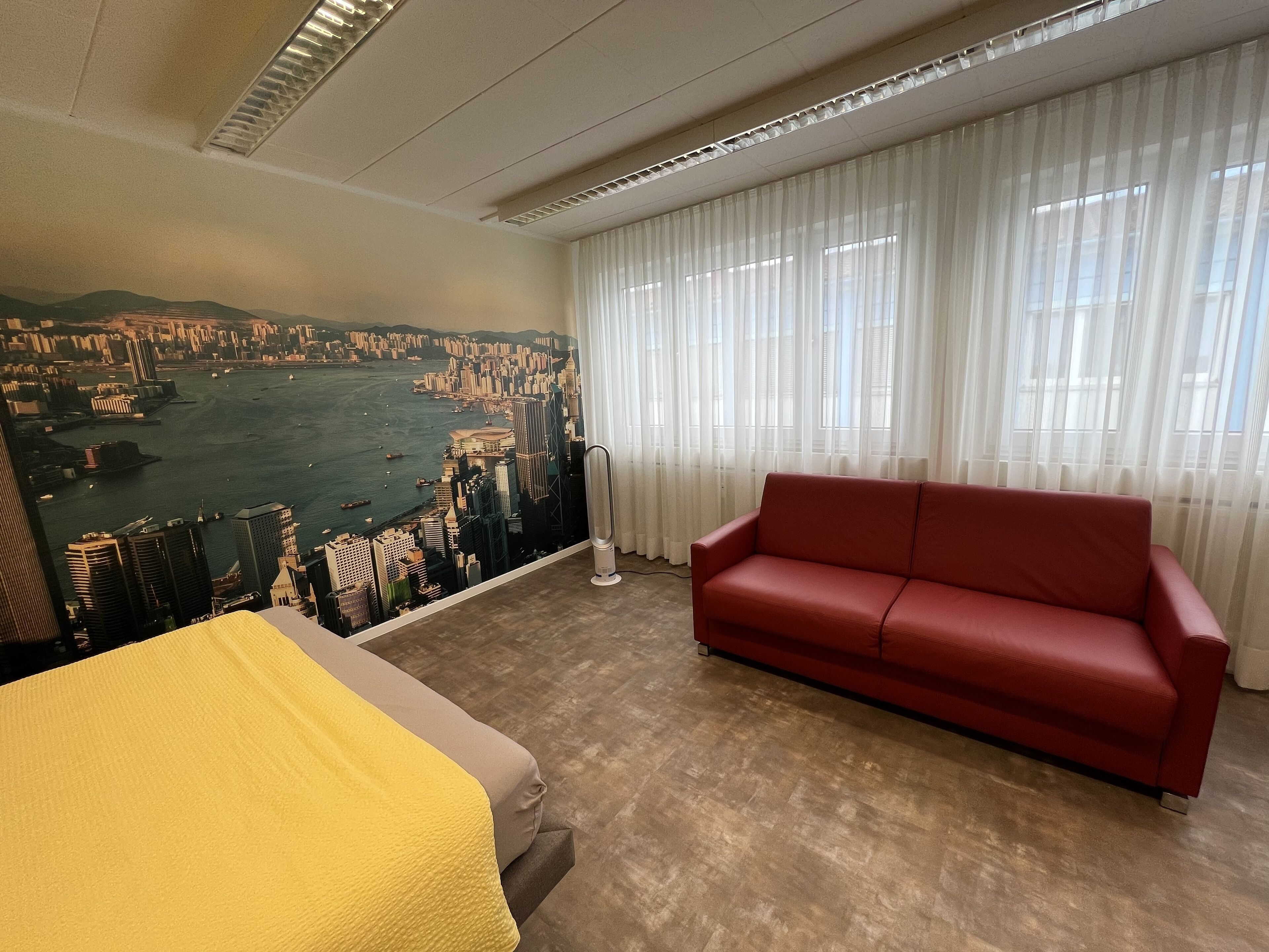 A hotel room with a large cityscape mural, a red sofa, a bed with a yellow blanket, and a view of a body of water through sheer curtains.