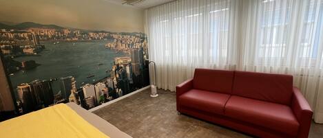 A hotel room with a large cityscape mural, a red sofa, a bed with a yellow blanket, and a view of a body of water through sheer curtains.