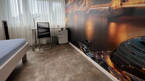 Habitació d'hotel amb llit, escriptori, cadira, paperera petita i finestra gran amb cortines de tul.