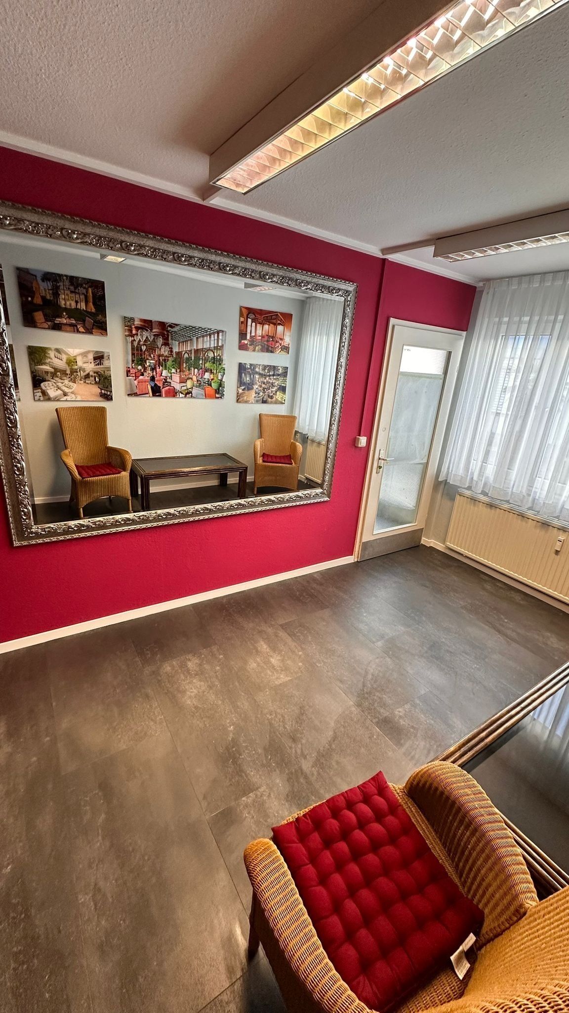 A spacious room with a large mirror, red walls, and a wooden floor. There are two wicker chairs and a red cushioned ottoman.