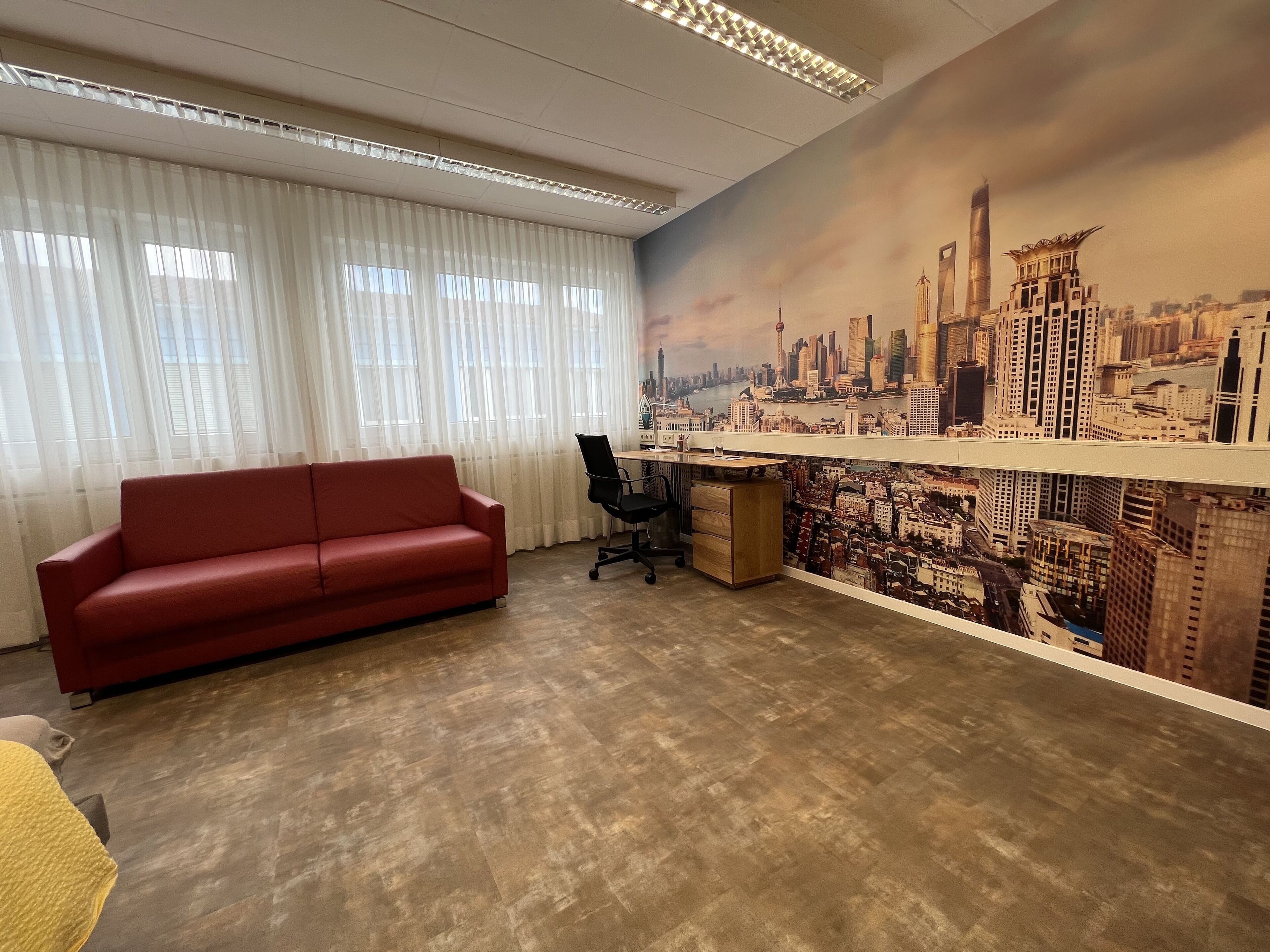 Een kamer met een rode bank, een houten bureau en een stadsgezicht muurschildering.